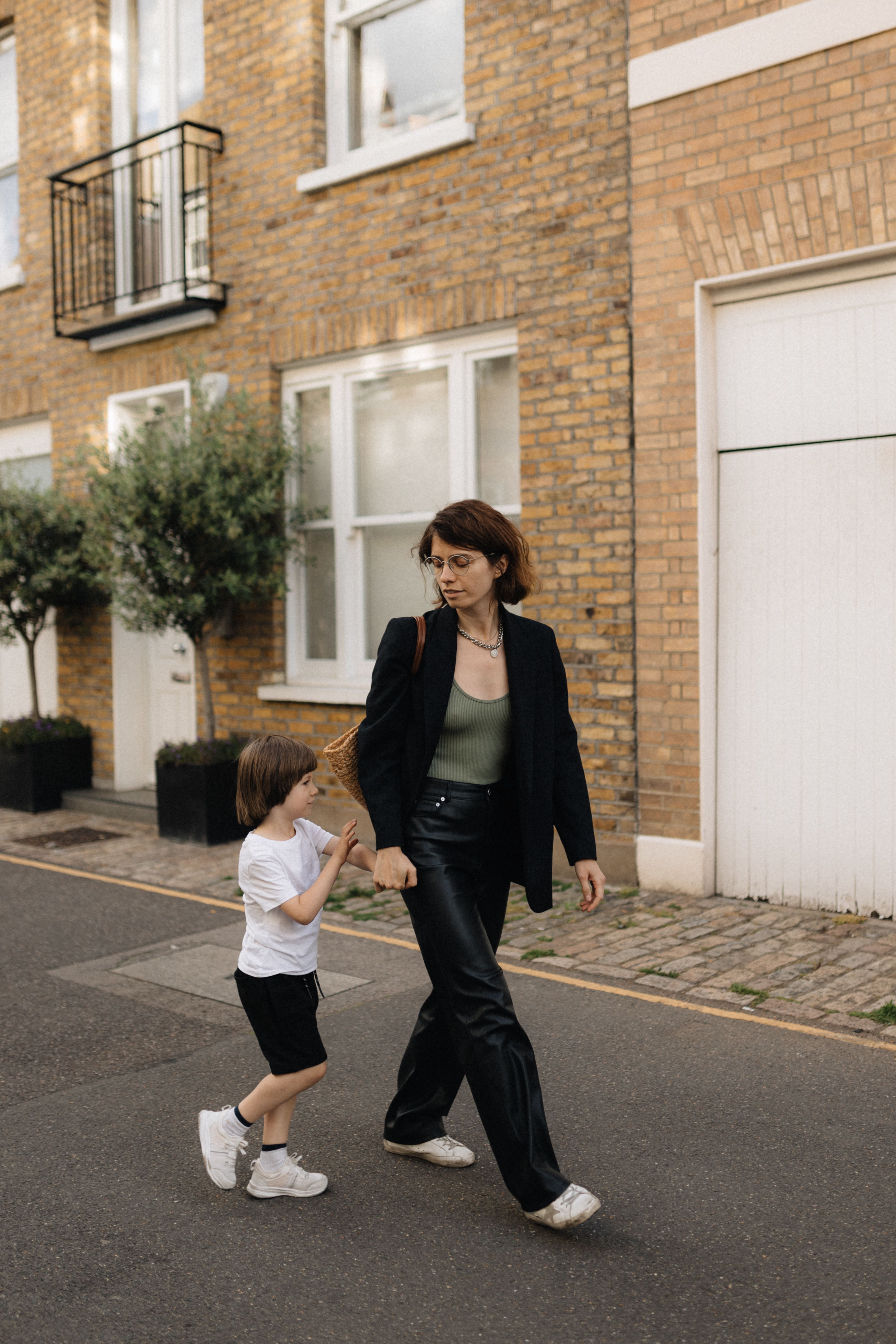 Photo of Ukrainian refugee Alisa Cooper and her son Kupriian. She is looking down at him as they walk through a terraced street in London. She has short brown hair and is wearing a black blazer and a green vest top.