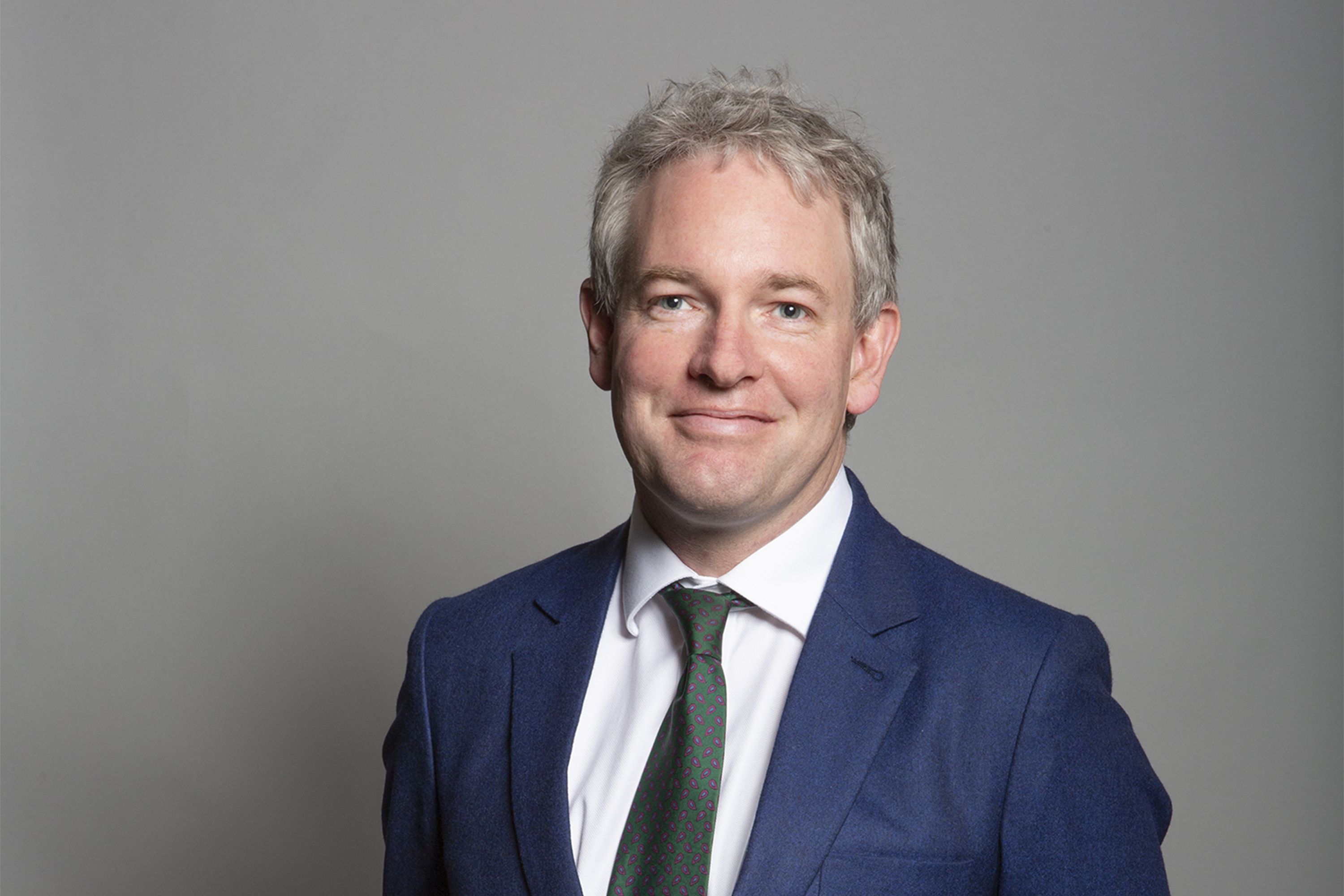 Portrait of Danny Kruger MP, a man with light grey hair wearing a navy blue suit jacket, white shirt and green tie.