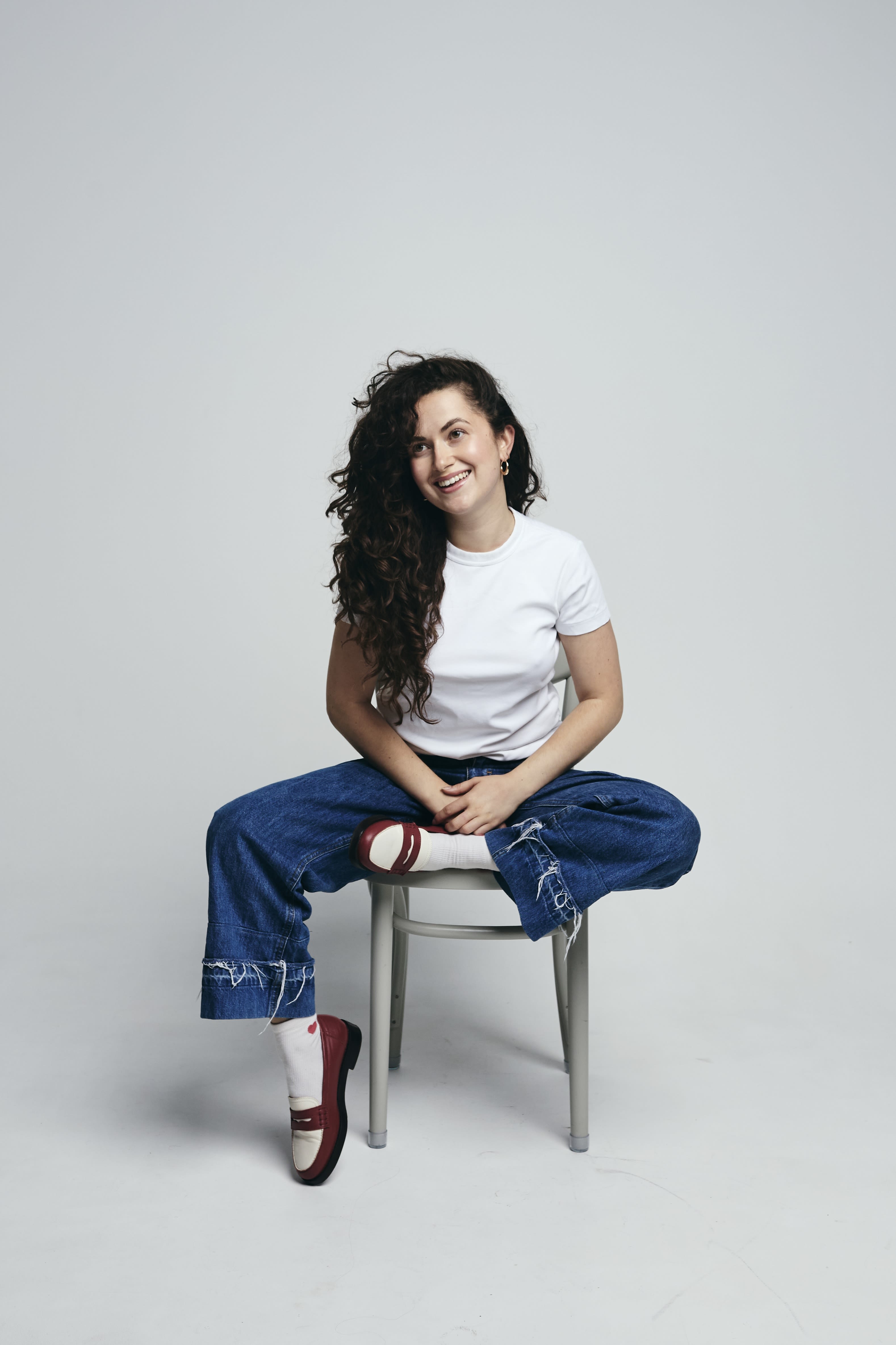 Olivia is sat on a stool against a white background, smiling. She is wearing loafers, jeans, a white t-shirt and has long curly hair