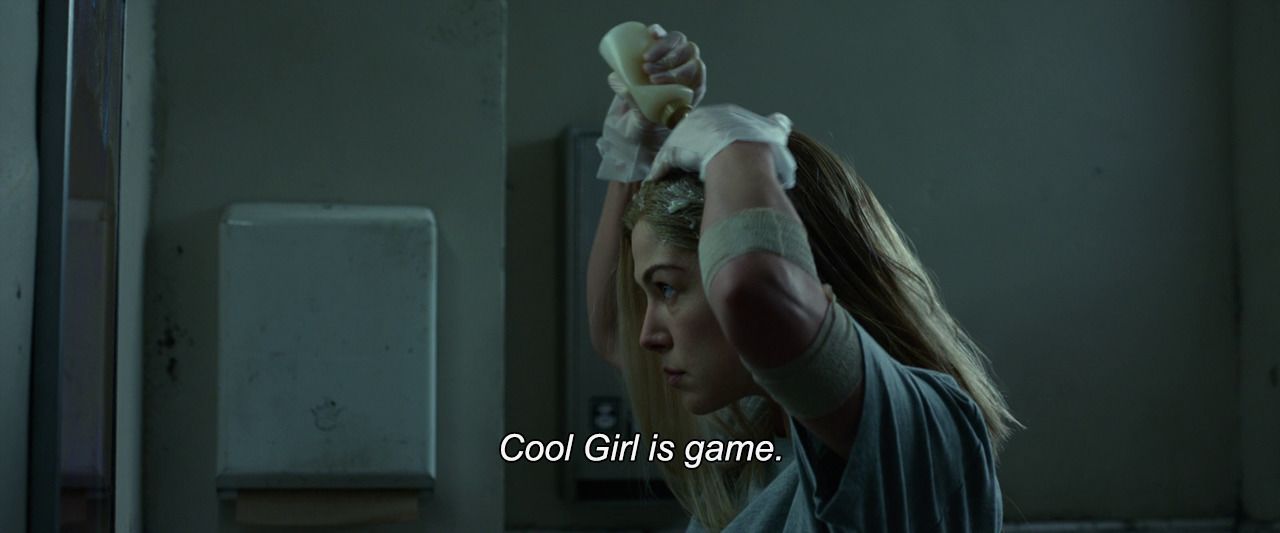 Rosamund Pike looks into mirror with one arm raised as she dyes her hair and the other arm on her head