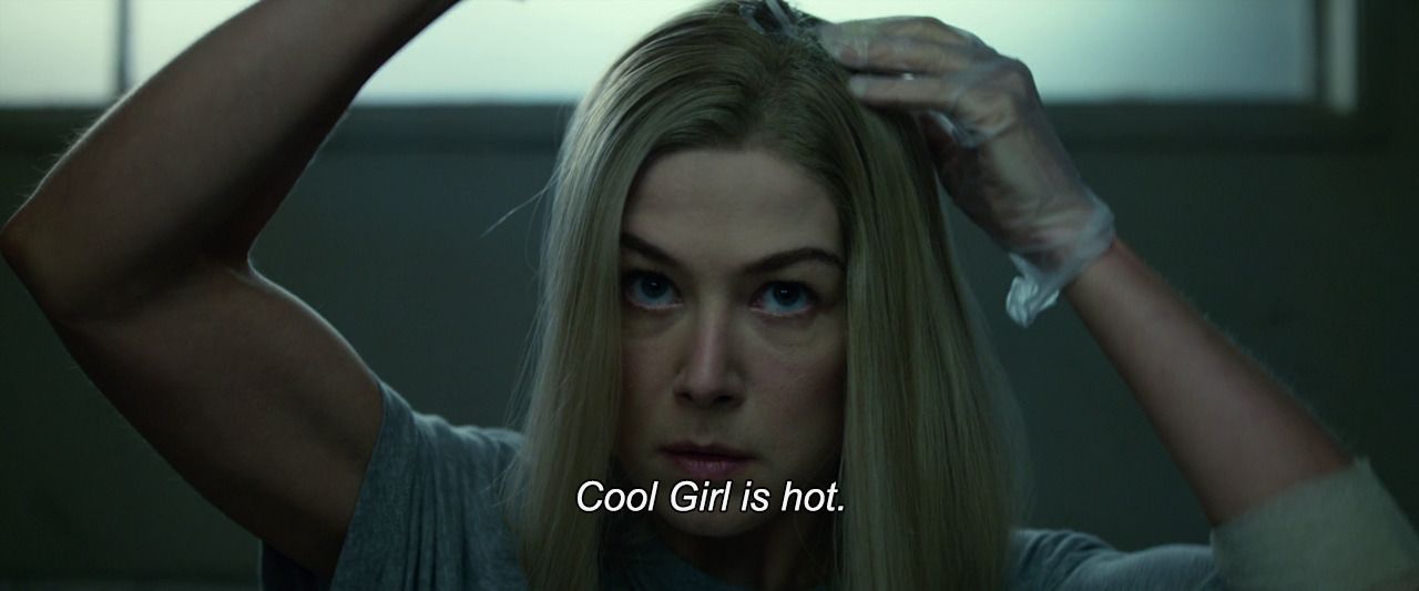 Rosamund Pike looks into camera with one arm raised as she dyes her hair and the other arm on her head