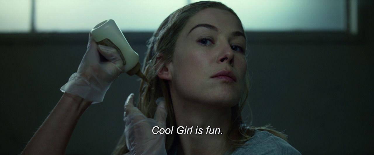 Rosamund Pike looks into camera with one arm raised as she dyes her hair and the other arm on her head