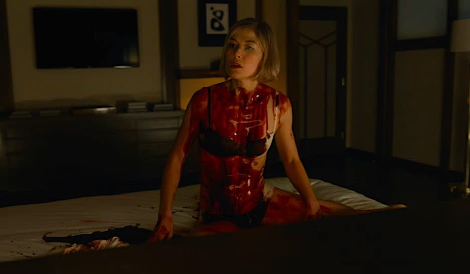 Rosamund Pike in underwear, covered in blood, sat on a bed, staring slightly left of camera
