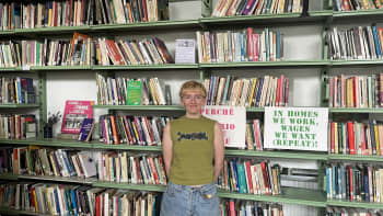 Woman with short blonde hair and piercings wearing a green vest and jean shorts stands in front of a colour bookshelf