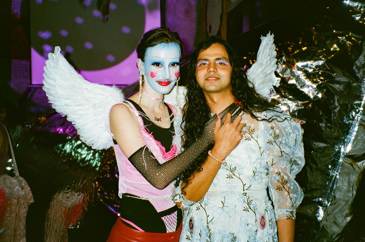 A man with face paint and angle wings embraces his partner wearing a dress