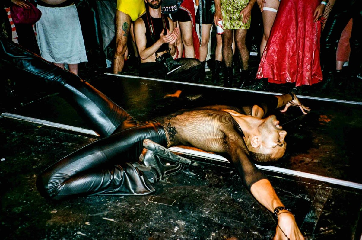 A man in leather trousers and a fishnet top "death dropping" on the floor in front of a crowd