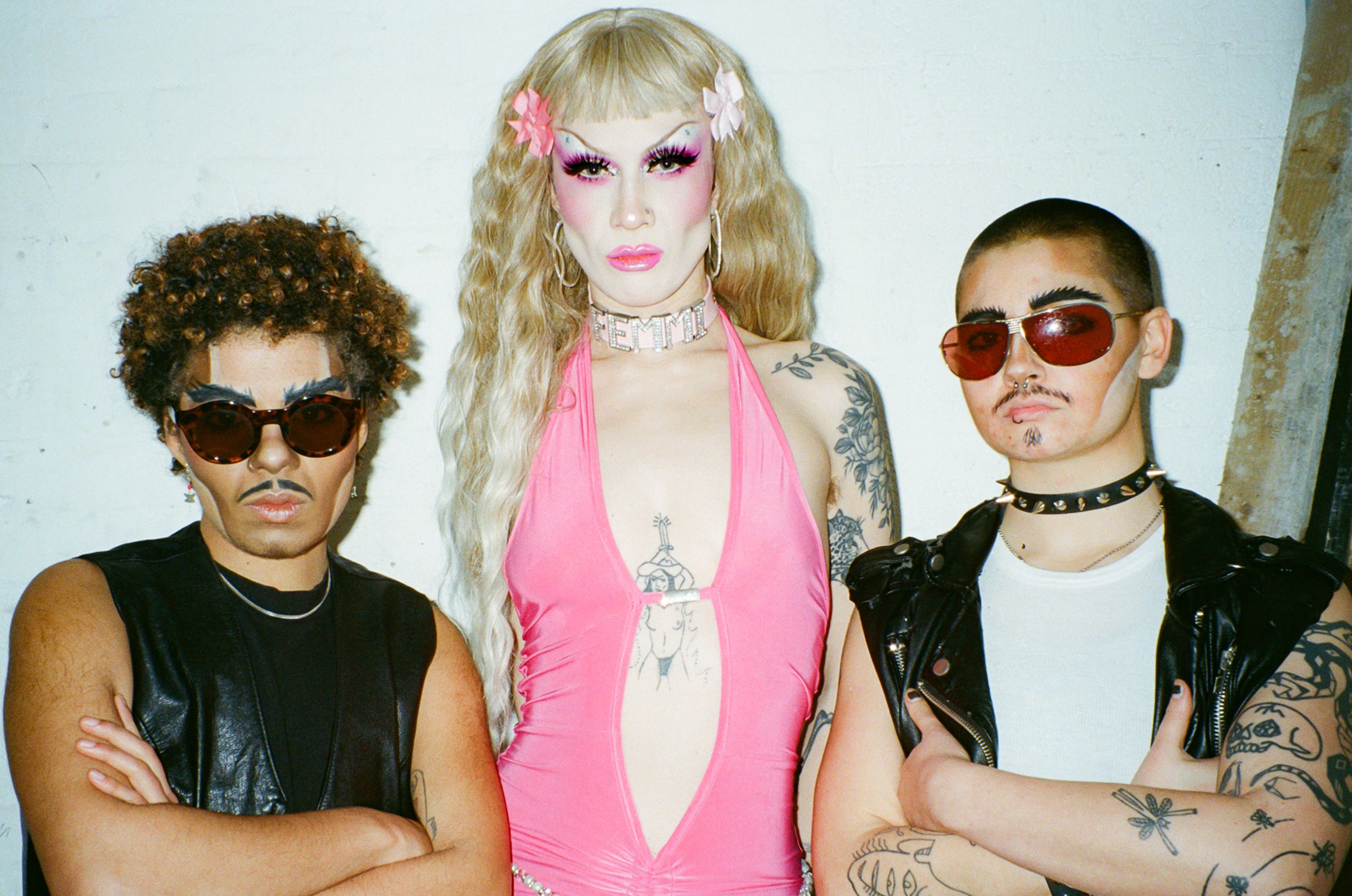 A woman with long blonde hair, drag makeup and a diamonte choker stands in between two women in male drag