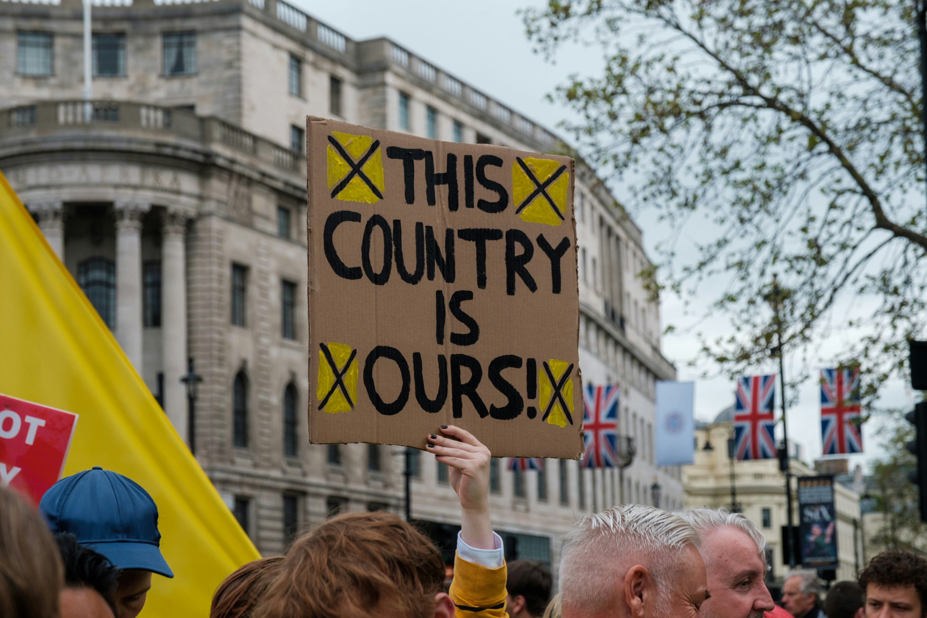 Protest scene in central London. Union Jack flags hang in the background. A protestor holds a cardboard sign that says 'this country is ours'