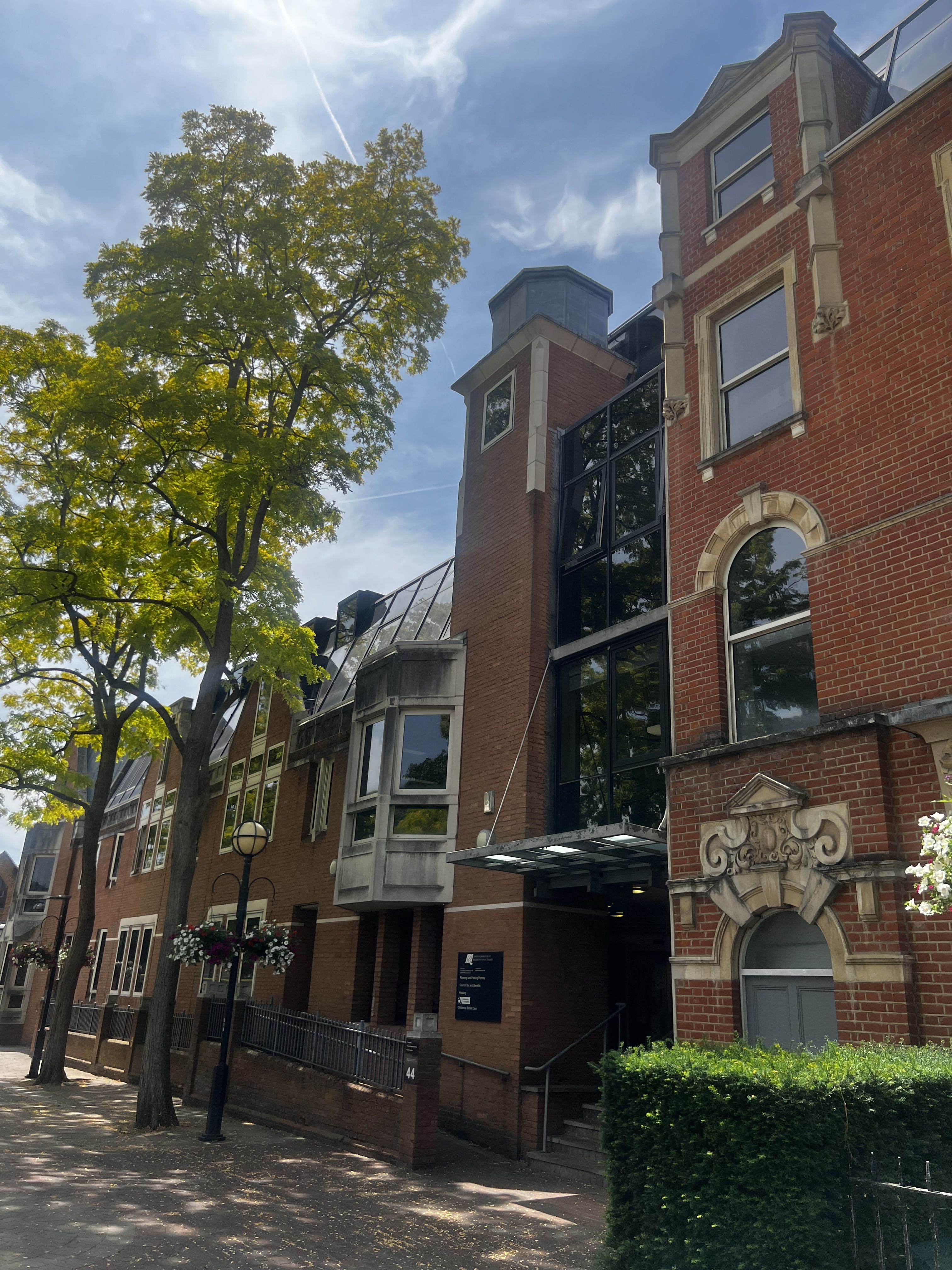 A large brick building featuring numerous windows across its facade, facing a set of trees to the left and bright blue sky.