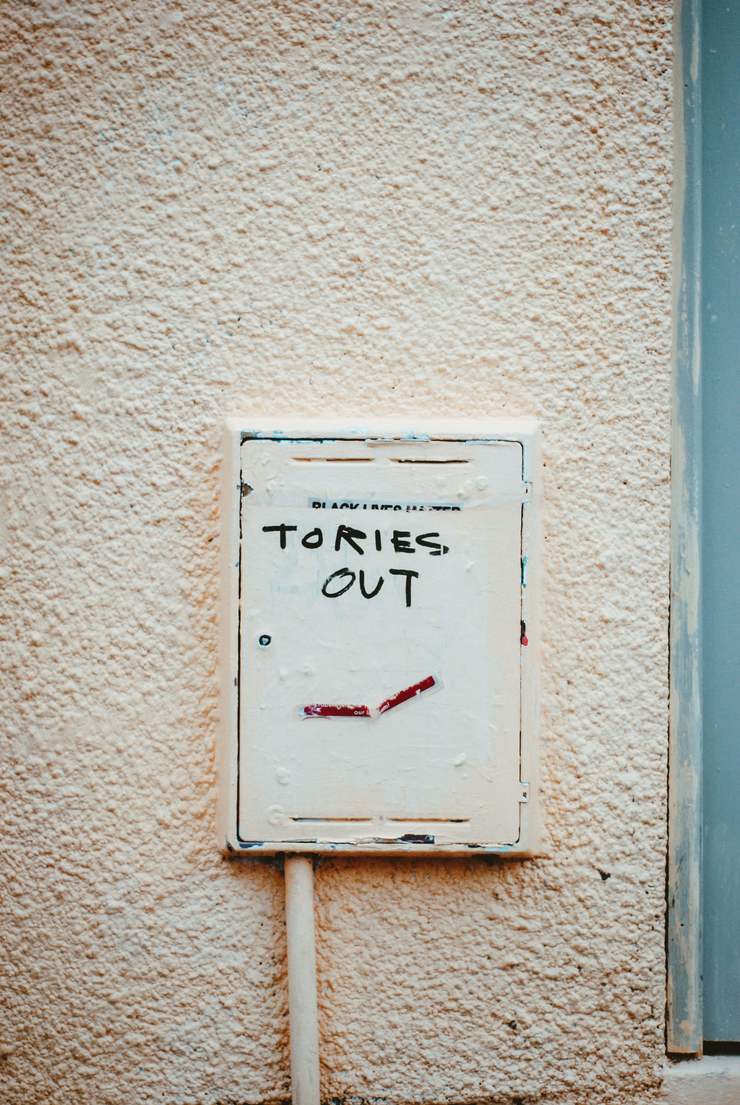 White speckled background of outside house wall, white electrical box central with black graffiti that reads "Tories out"